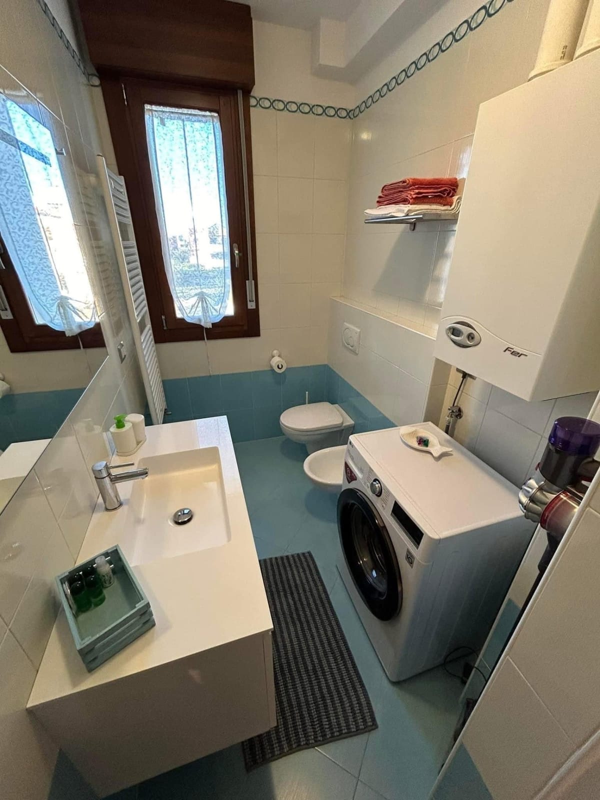 Bagno con piastrelle turchesi, lavabo, sanitari, lavatrice e finestra